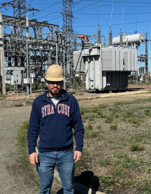 Man in Syracuse sweatshirt and hard hat stands in front of a power substation