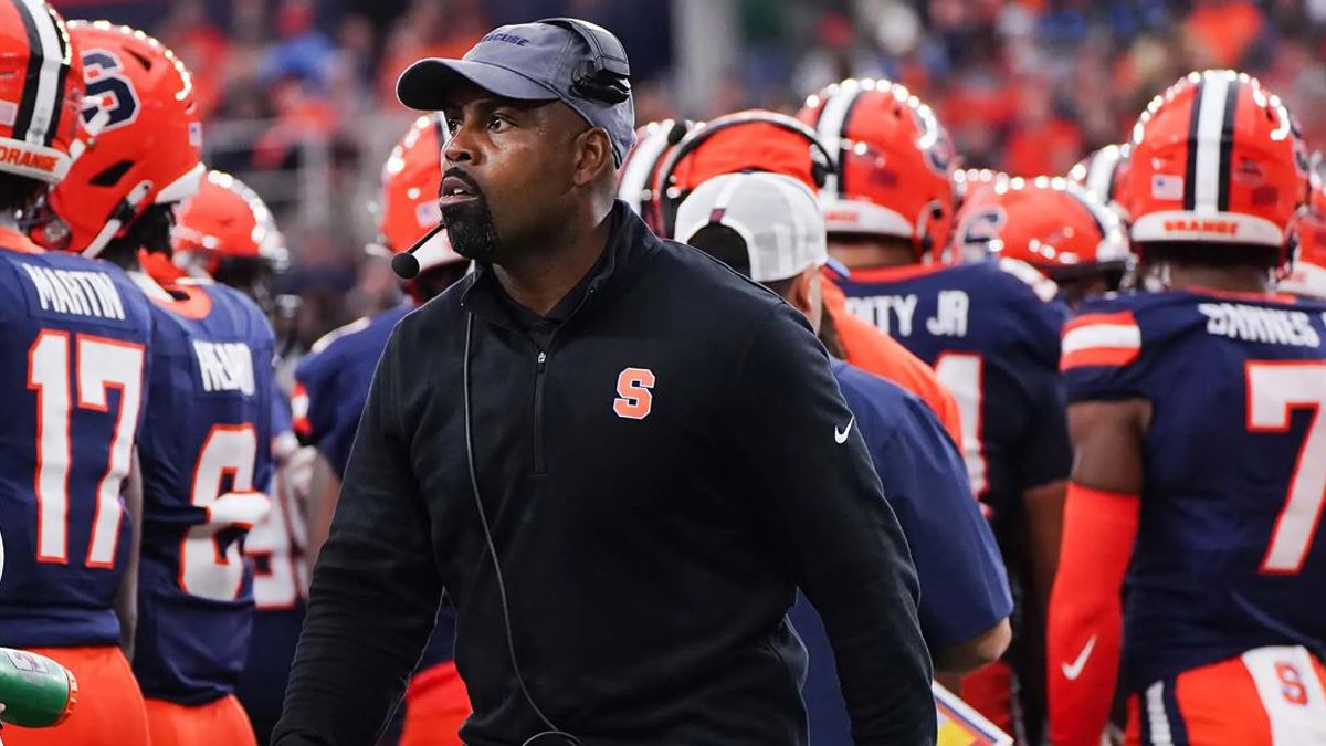 A football coach walks up the sideline while surrounded by his players.