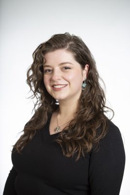 Smiling person with long wavy hair, wearing a black top and turquoise earrings, posed against a plain white background.