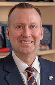 A person in a dark blue suit, white shirt and blue and orange striped tie stands in front of a bookshelf with framed photos and books.