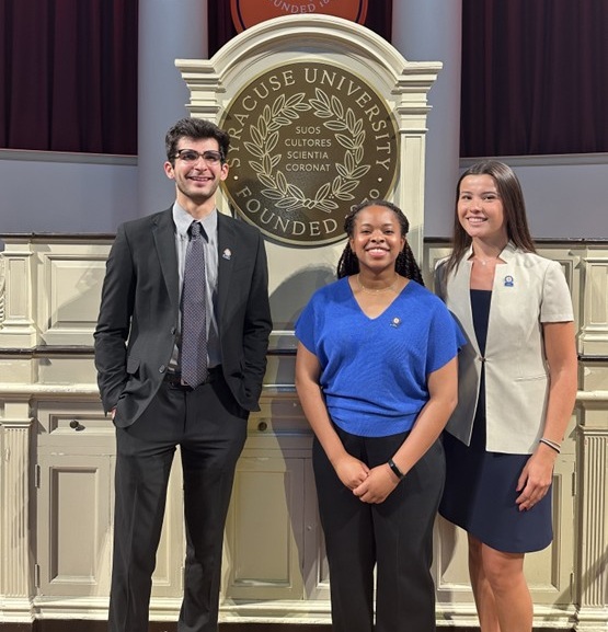 Three individuals standing in front of a podium with the Syracuse University emblem, which reads 'Syracuse University Founded 1870' and includes the Latin phrase 'Suos Cultores Scientia Coronat.' A red banner with the same emblem hangs above the podium.
