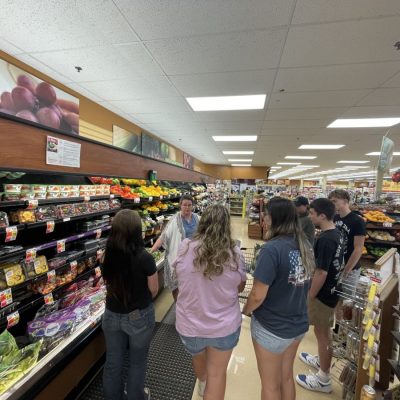 Group of people standing in the produce section of a grocery store, listening to a speaker. Shelves are stocked with fresh fruits and vegetables including packaged grapes and bell peppers, suggesting an educational or informational activity.