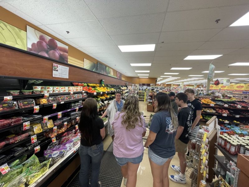 Group of people standing in the produce section of a grocery store, listening to a speaker. Shelves are stocked with fresh fruits and vegetables including packaged grapes and bell peppers, suggesting an educational or informational activity.