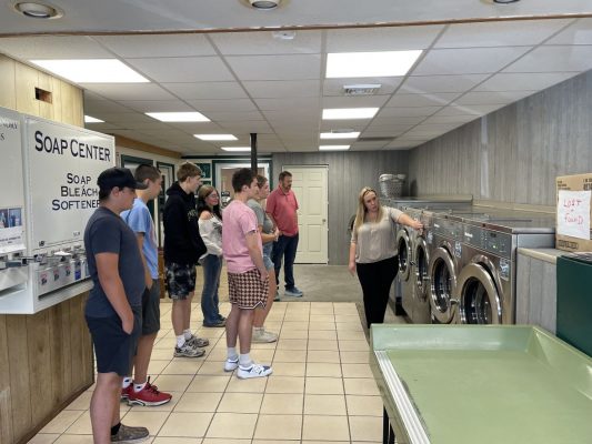 Group of people standing in a clean, well-lit laundromat with tiled flooring. One person is pointing at a washing machine, possibly explaining something to the group. A 'SOAP CENTER' sign on the wall reads 'SOAP BLEACH SOFTENER – $1.25 each,' and multiple washing machines are lined up along the wall.
