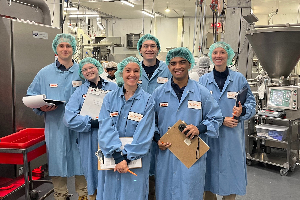 Group of seven people in blue lab coats and hair nets holding clipboards and smiling in an industrial food processing facility with stainless steel equipment.