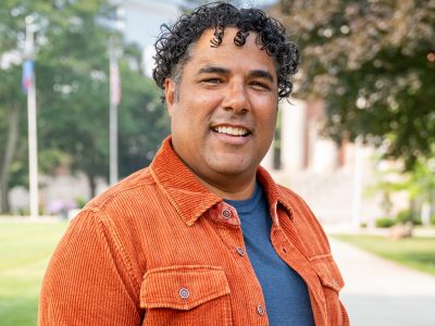 Person with curly hair in an orange corduroy jacket and blue shirt, standing outdoors with trees and a building in the background