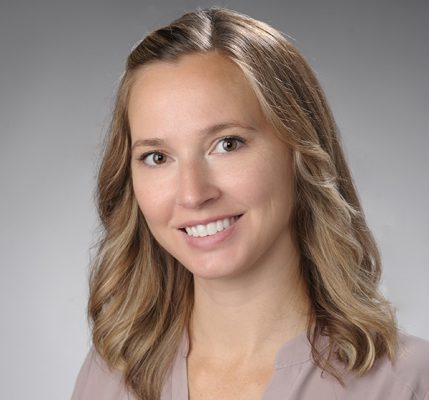 A faculty member smiles while posing for a headshot in front of a grey backdrop.
