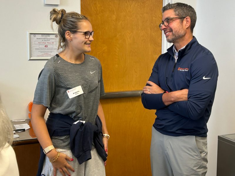 Two individuals, both wearing glasses, converse casually indoors near a wooden door; one wears a gray Nike t-shirt, the other a navy quarter-zip pullover and light gray pants.