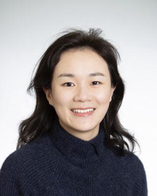 Person with shoulder-length dark hair, wearing a dark blue sweater, smiling against a plain white background.
