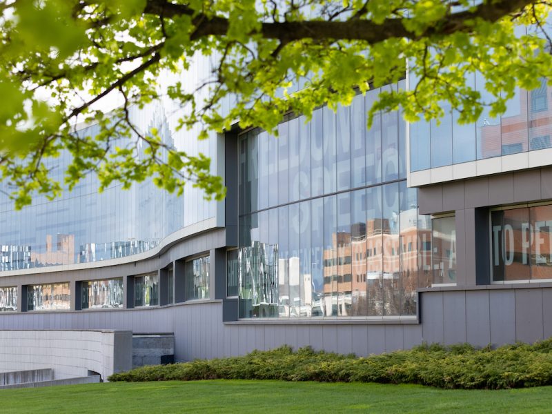 Modern glass building on Syracuse University campus framed by trees and greenery.