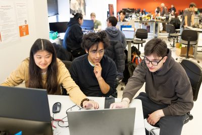 Team of three individuals collaborating around a laptop in a classroom, with others working in the background.