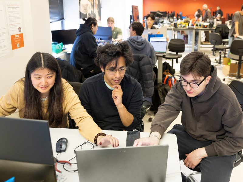 Team of three individuals collaborating around a laptop in a classroom, with others working in the background.