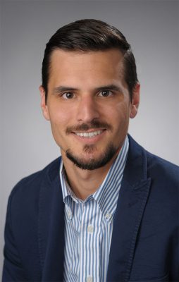 Person with short dark hair and closely trimmed beard and mustache wearing a dark blue jacket and a white and light blue striped shirt