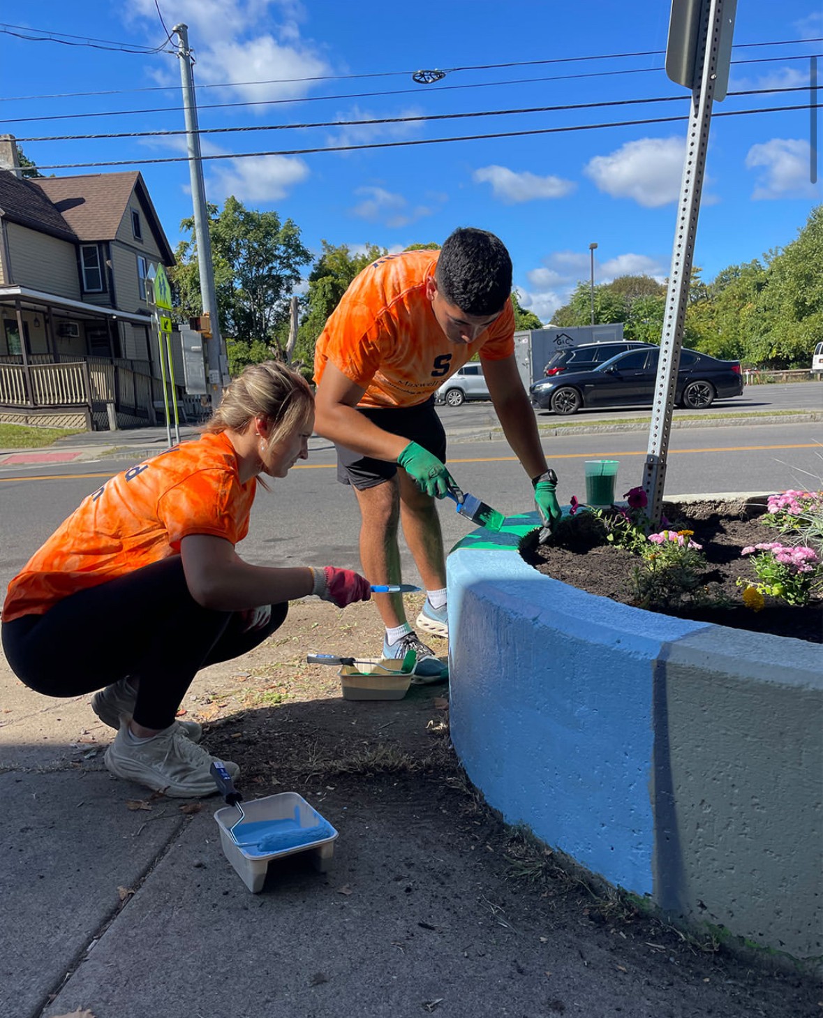 Two volunteers in orange shirts painting a blue concrete planter and planting flowers on a city street, with paint supplies nearby and residential buildings in the background