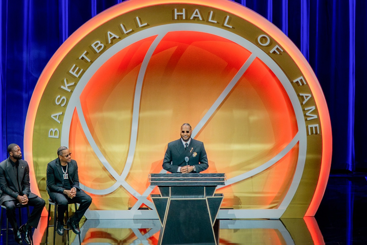A speaker stands at a podium on stage at the Basketball Hall of Fame, with the organization's distinctive circular logo featuring a basketball design prominently displayed behind them.