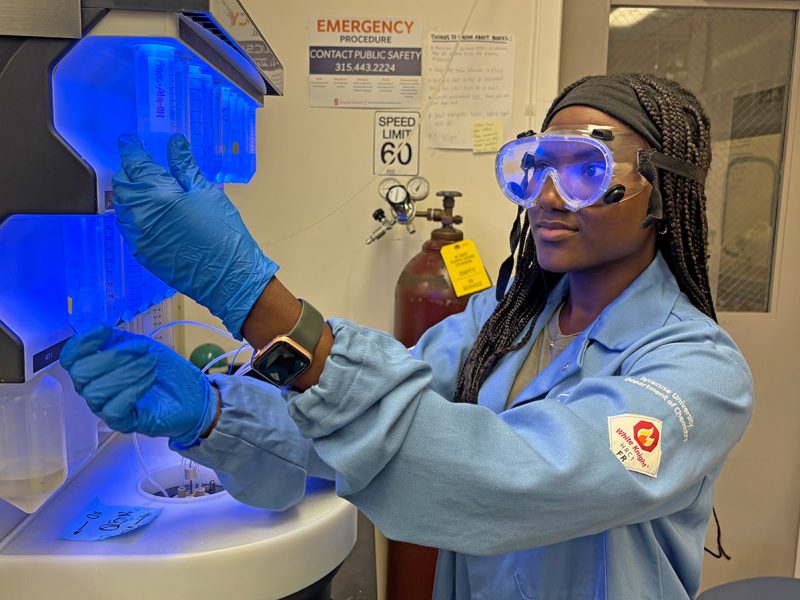 A woman wearing safety goggles, blue gloves, and a light blue lab coat works with a glowing blue laboratory instrument in a research lab