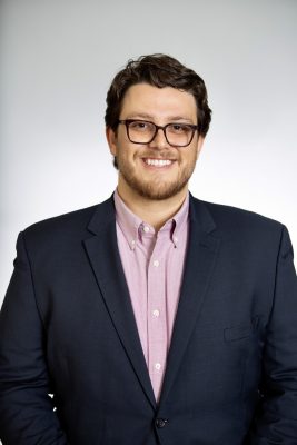 Person wearing glasses, dark blazer and light pink shirt, smiling against a plain white background