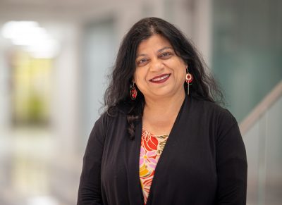 Person with long dark hair wearing a black top over a colorful patterned shirt and red-gold earrings, standing in an indoor setting with a blurred background