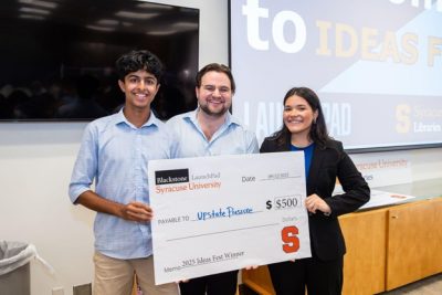 Three people pose with a large $500 check from Blackstone LaunchPad at Syracuse University, awarded to "Upstate Rescue" as the 2025 Ideas Fest Winner, with a projection screen visible in the background.