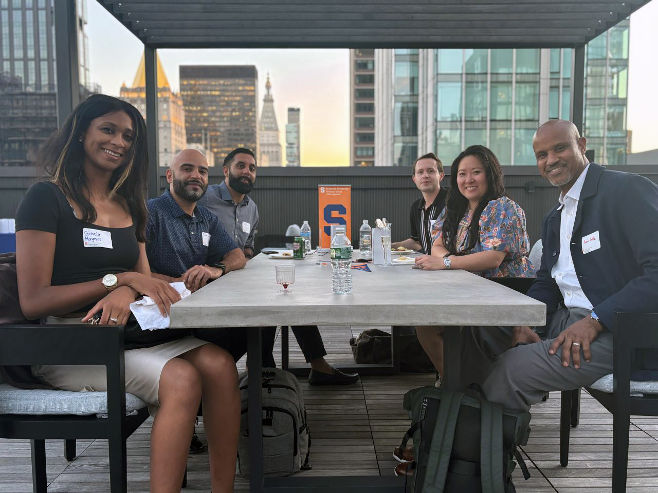 Six people seated around a rooftop table during a professional networking event, with city skyline in the background