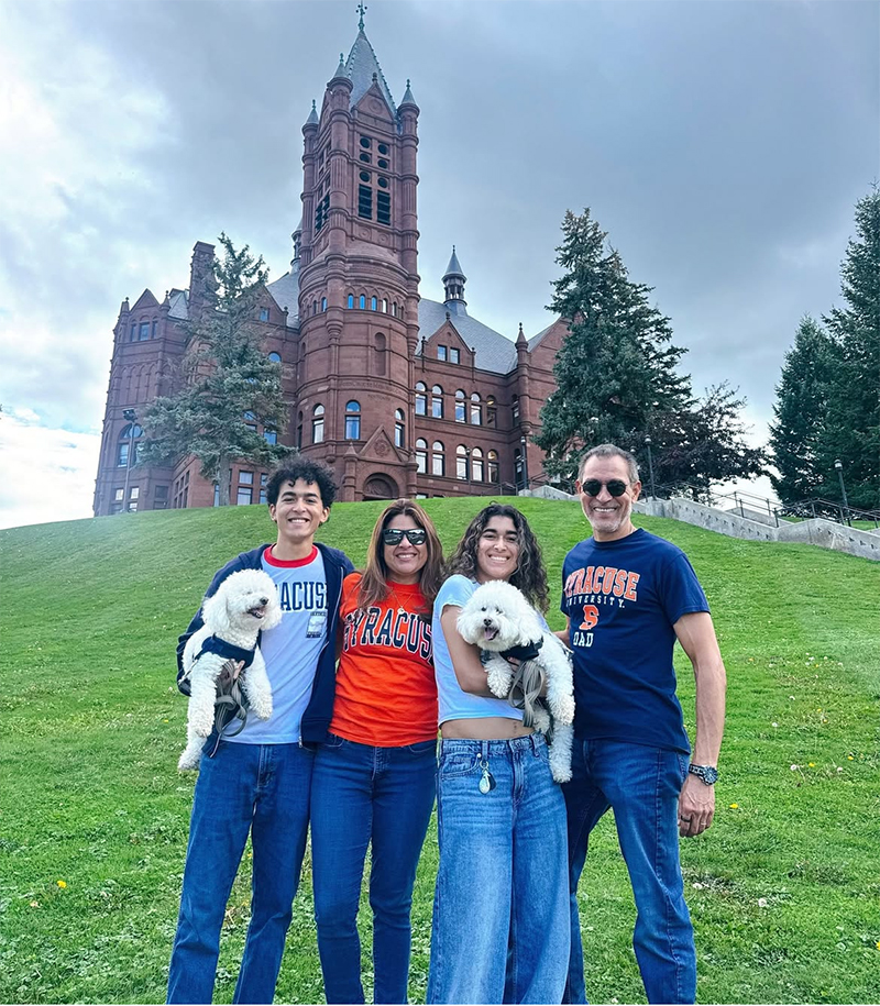 four people in Syracuse University apparel holding two small white dogs, stand on a grassy hill in front of a historic red-brick campus building