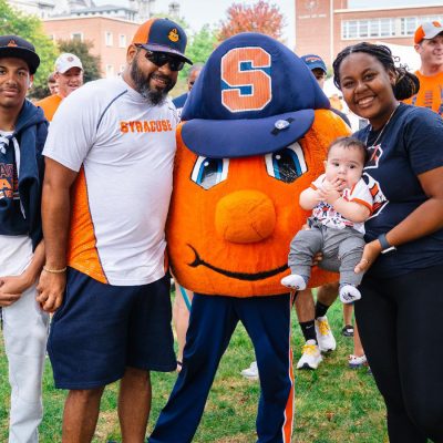Group photo with Syracuse University mascot Otto the Orange on campus lawn, surrounded by people in Syracuse apparel, including a baby in themed clothing.