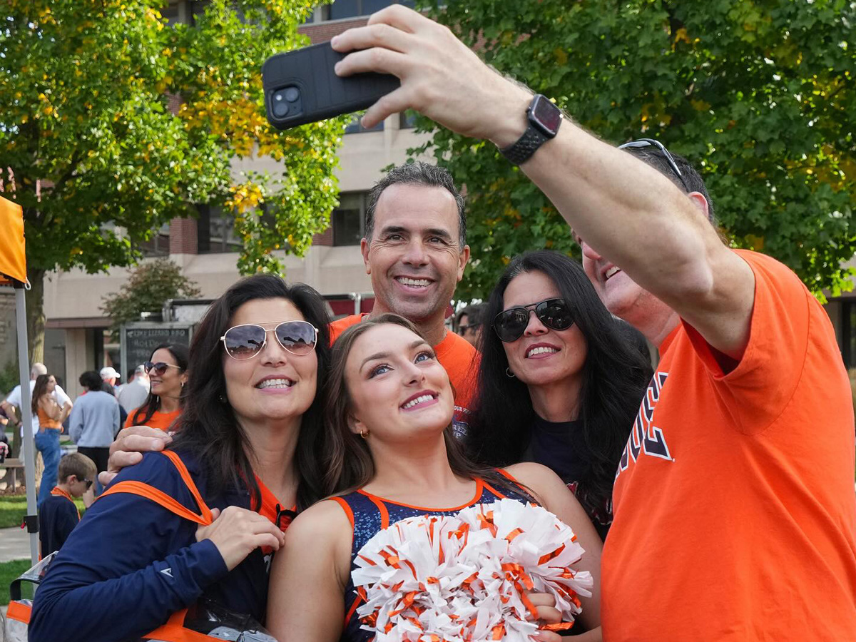 Group selfie of five people, including a cheerleader with pom-poms, wearing Syracuse-themed orange and blue attire in an outdoor campus setting