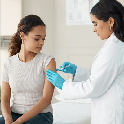 Healthcare professional administering a vaccine to a seated person in a clinical exam room.