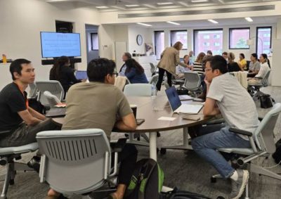 Participants seated around tables in a classroom or conference room, engaging in discussion and note-taking during the Course Redesign Institute. A large screen at the front displays the event title, and the room is well-lit with natural light from several windows.