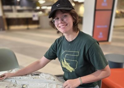 Person in a black cap and green graphic t-shirt sits indoors, examining a light jacket on a table, with blurred furniture and decor in the background