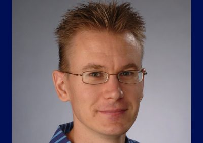 Professional headshot of a man with short spiky blonde hair wearing wire-rimmed glasses and a blue striped button-down shirt against a gray background.
