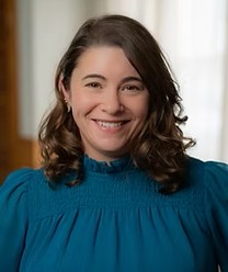 A person with side parted curly brown shoulder length hair wearing a teal blue top