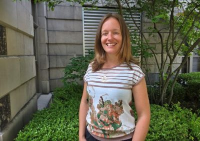Person with shoulder-length brown hair wearing a striped floral shirt stands in front of a concrete wall and outdoor space.
