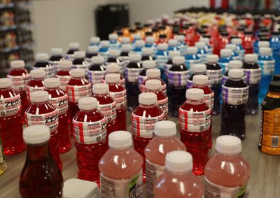 Rows of colorful Powerade Zero bottles in assorted flavors displayed on a table, with white caps and visible branding; background includes shelves and additional bottles.